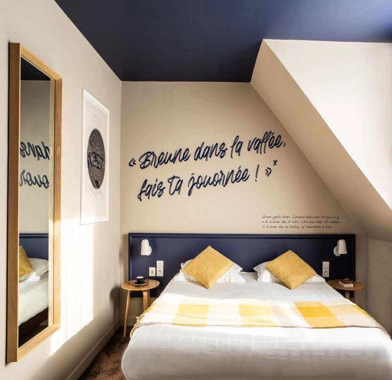 Chambre cosy avec tête de lit bleue, coussins jaunes et citation décorative murale à l’Hôtel Antares & Spa Honfleur.