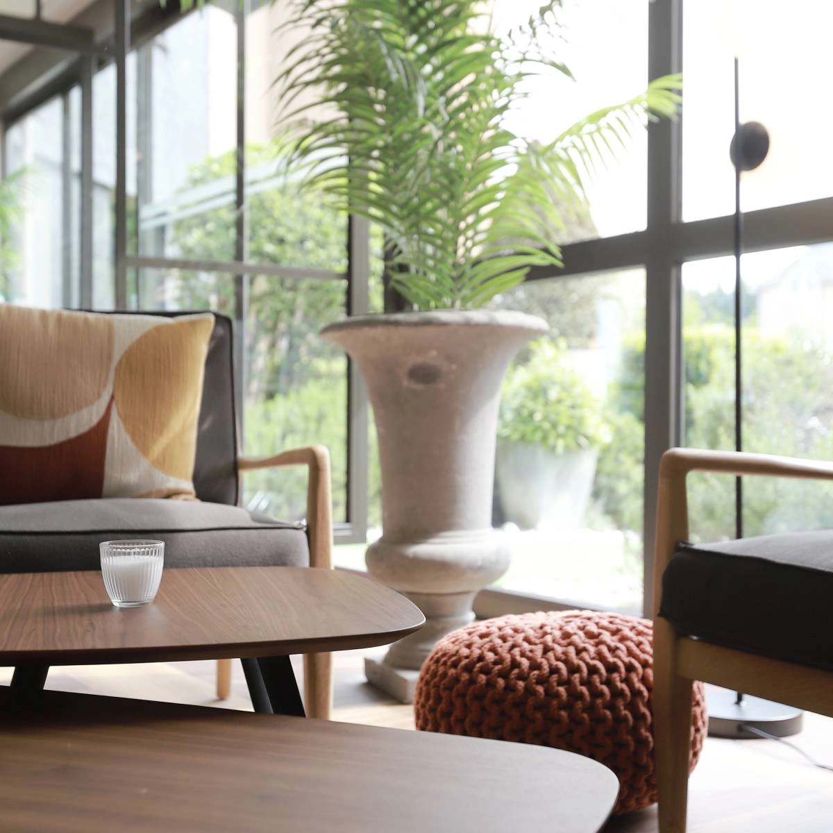 Espace détente avec fauteuil, table basse, pouf tressé et grande plante près d’une baie vitrée à l’Hôtel Antares & Spa Honfleur.