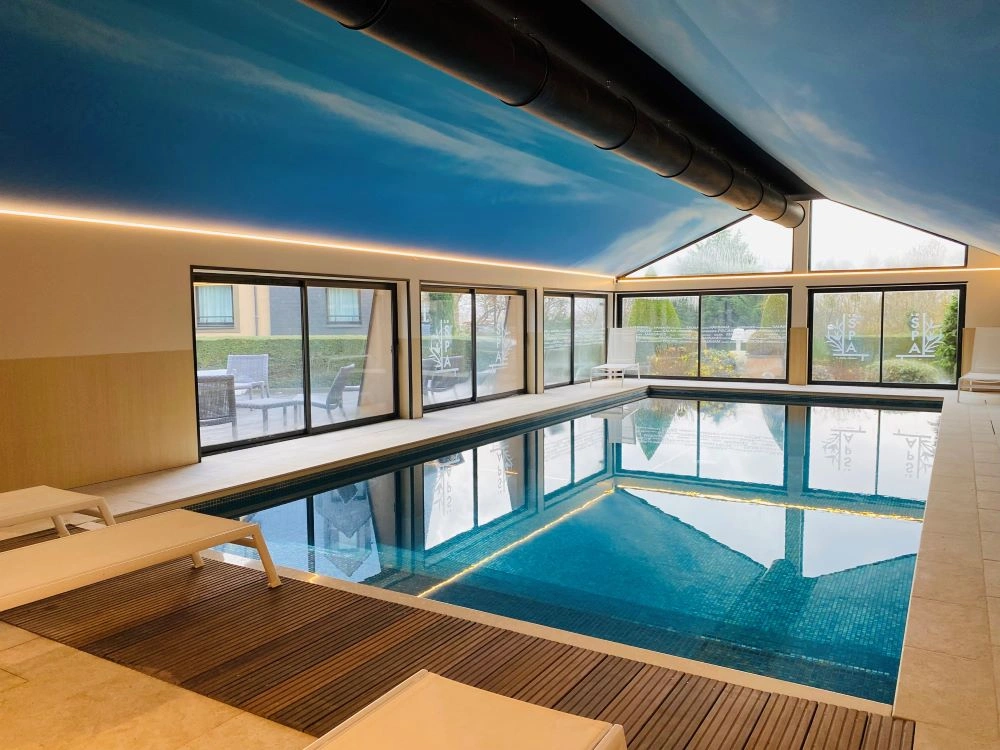 Piscine intérieure avec transats, grandes baies vitrées et plafond bleu à l’Hôtel Antares & Spa Honfleur.